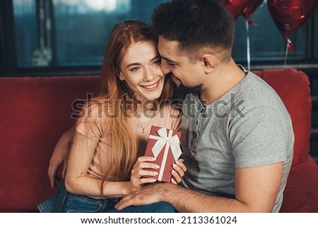 Caucasian couple, girl holding in hands red gift box for anniversary congrats sitting on sofa. Happy birthday. Celebrate party. Valentines day. Helium balloons.