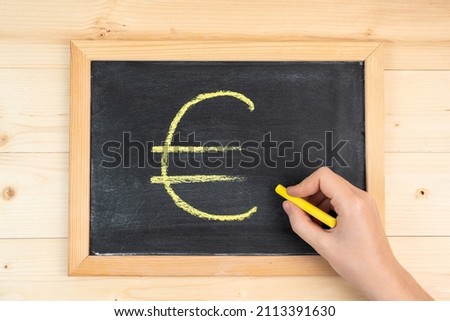 A woman's hand draws the euro symbol in yellow chalk on a black blackboard in a frame