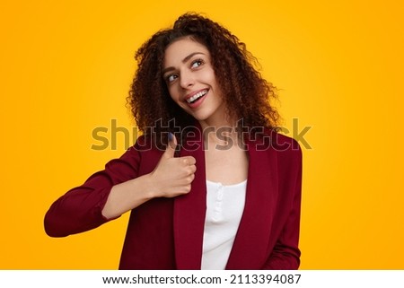 Optimistic young female with curly hair showing like gesture and looking up dreamily against yellow background