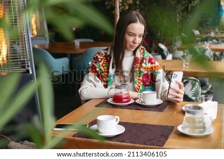 A young brunette girl is sitting in a cafe drinking tea, resting, talking on the phone, taking pictures, recording reels videos for social networks. Positive emotions. The view behind the glass.