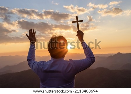 silhouette of man praying and holding christian cross for worshipping God at golden color sunset background,.Christian, Christianity, Religion copy space background.