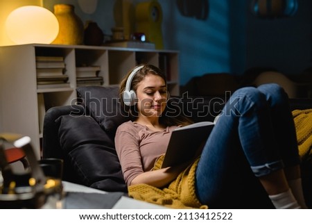 woman relaxing on a sofa at home. listening music on headphones and reading book Royalty-Free Stock Photo #2113412522