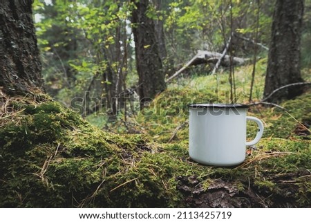Enamel white mug in the mossy wood mockup. Trekking merchandise and camping geer marketing photo. Stock wildwood photo with white metal cup. Rustic scene, product mockup template. Lifestyle outdoors.