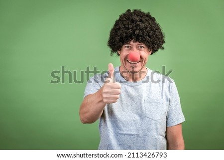 Man with black wig and red clown nose posing in front of green background