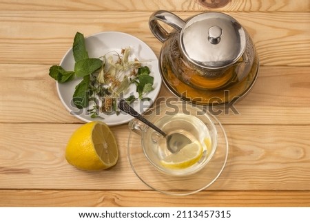 Herbal tea brewed in the glass teapot with strainer, same tea in the glass cup with spoon, herbs for tea preparation on saucer and lemon on rustic table, top view
