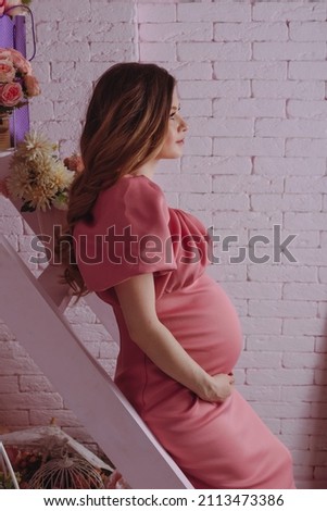Pregnant woman in pink dress sitting on the stairs furnished with bouquet of flowers