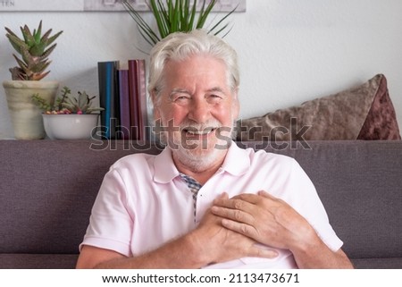 Happy thankful smiling old man holding hands on chest sitting on sofa at home feeling love and gratitude. Beautiful white haired grandfather wearing pink polo looking at camera,  peace of mind concept Royalty-Free Stock Photo #2113473671