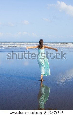 Young woman walking barefoot on empty beach. Full body portrait. Slim Caucasian woman wearing long dress. View from back. Water reflection. Freedom concept. Vacation in Asia. Bali, Indonesia
