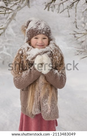a beautiful blonde girl is standing in a winter forest covered in snow dressed in a brown hat and coat with a large knitted scarf