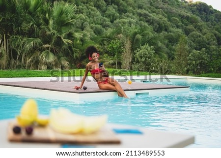 A cheerful young girl of African descent relaxes by tanning in the sun with her feet in the pool and listening to music with headphones - young African American woman drinks a healthy fruit cocktail
