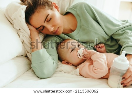 New mom taking a nap with a bottle of formula milk in her hand. Caring mom resting while her baby sucks on a pacifier. Mother and baby lying on the bed together. Royalty-Free Stock Photo #2113506062