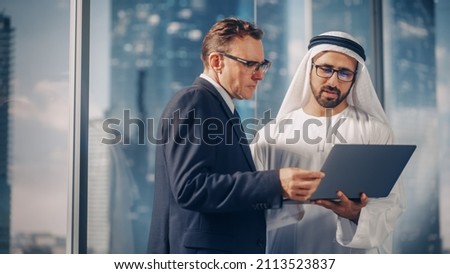 International Operations Manager Meeting Saudi Business Partner in Traditional Kandura. They're Standing in Modern Office, Using Laptop Computer. Successful Saudi, Emirati, Arab Businessman Concept. Royalty-Free Stock Photo #2113523837