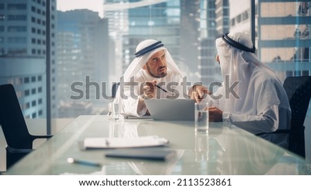 Two Successful Emirati Businessmen in White Traditional Kandura Sitting in Office Meeting, Negotiating and Talking About Financial Opportunities, Using Laptop. Saudi, Emirati, Arab Businessman Concept Royalty-Free Stock Photo #2113523861