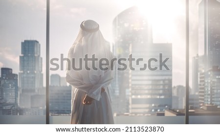 Successful Muslim Businessman in Traditional White Outfit Standing in His Modern Office Looking out of the Window on Big City with Skyscrapers. Successful Saudi, Emirati, Arab Businessman Concept. Royalty-Free Stock Photo #2113523870