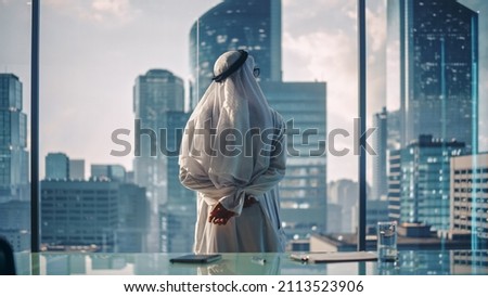 Successful Muslim Businessman in Traditional White Kandura Standing in His Modern Office Looking out of the Window on Big City with Skyscrapers. Successful Saudi, Emirati, Arab Businessman Concept. Royalty-Free Stock Photo #2113523906