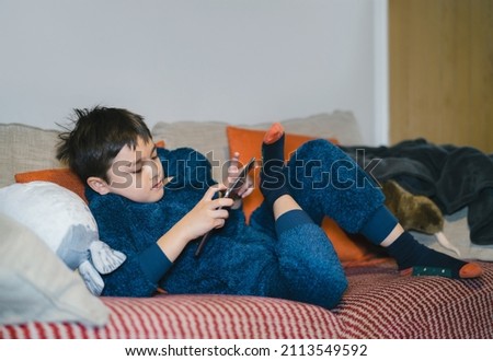Cinematic portrait happy kid watching cartoon on tablet, Candid shot cute boy playing game online with friends on internet, Cozy Child wearing fluffy pajamas sitting on sofa relaxing at home 