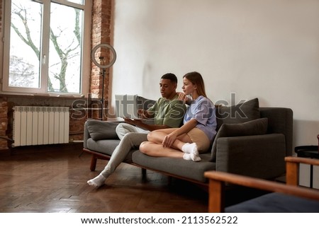 Young multiracial blogger couple editing video content on laptop for social networks. Concept of video blogging and internet multimedia. Caucasian girl and black guy of generation Z on sofa at home
