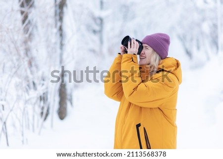 Cheerful girl photographer in a yellow jacket takes pictures of winter in a snowy park
