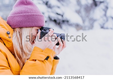 Cheerful girl photographer in a yellow jacket takes pictures of winter in a snowy park
