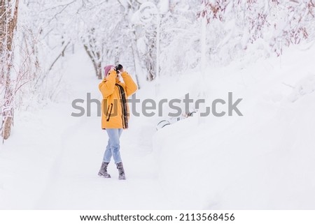 Positive girl in yellow jacket photographer taking pictures of snow in winter park