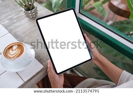 Top view mockup image of a woman holding digital tablet with blank white desktop screen with coffee cups on the table