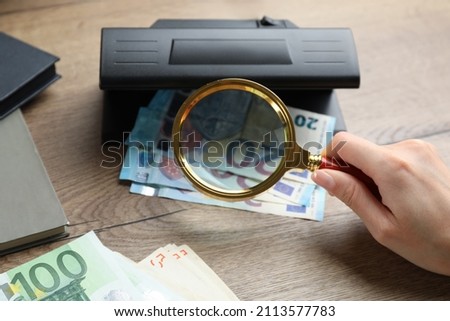 Woman checking Euro banknotes with currency detector and magnifying glass at wooden table, closeup. Money examination device