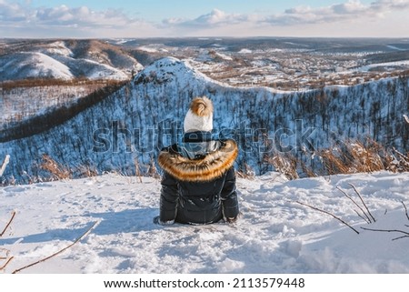 A happy little girl in warm clothes stands on a mountain with a beautiful winter snowy landscape