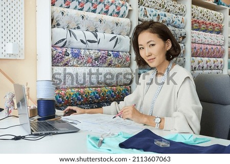 Pretty Asian woman fashion designer working on laptop in workshop, sketching clothes models, smiling, looking at camera