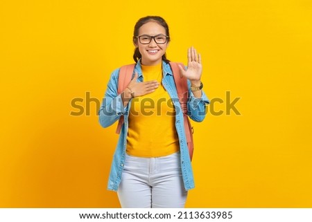 Smiling young Asian woman student in denim outfit with backpack with hands on chest and waving to camera isolated on yellow background. Education in university college concept