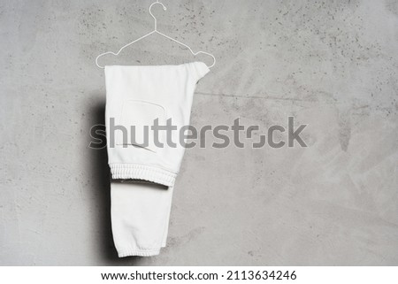 Mockup of blank white sweatpants hanging on the thin metallic hanger against light concrete wall