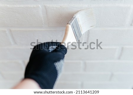 Close-up of a worker's hand with a brush painting a brick wall with white paint.