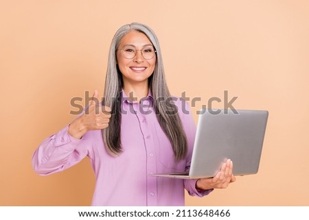 Photo of funny white hairdo elder lady hold laptop show thumb up wear eyewear purple shirt isolated on beige background