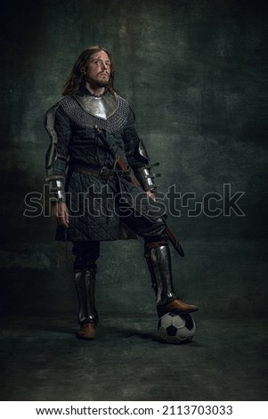 Ancient soccer player. Portrait of medieval warrior or knight with dirty wounded face holding isolated over dark background. Comparison of eras, history, renaissance style. Fashion, beauty, vintage Royalty-Free Stock Photo #2113703033