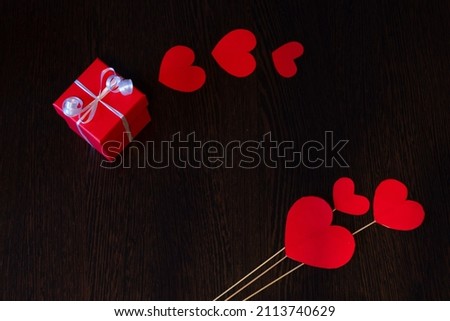 A gift packed in a red box and tied with a white ribbon, on a dark background. A gift, surrounded by red hearts made of cardboard, on a dark background.