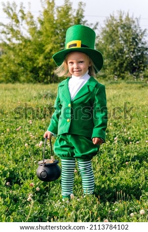 A girl in a leprechaun costume holds a katelok with dollar coins in her hand. The Toddler child stands in a clearing of green clover and smiles happily. St. Patrick's Day Holiday