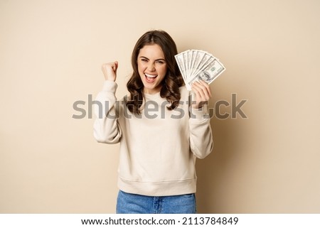 Enthusiastic modern woman winning money, got cash, celebrating and shouting of joy, standing against beige background