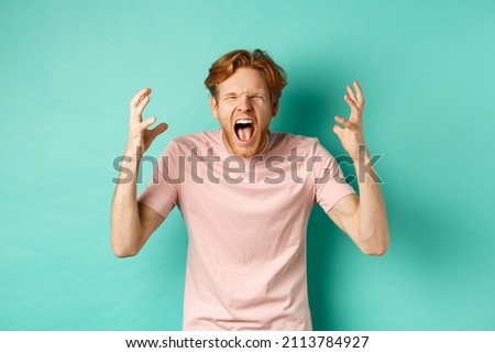Angry young redhead man screaming distressed, shaking hands and looking outraged, standing over turquoise background