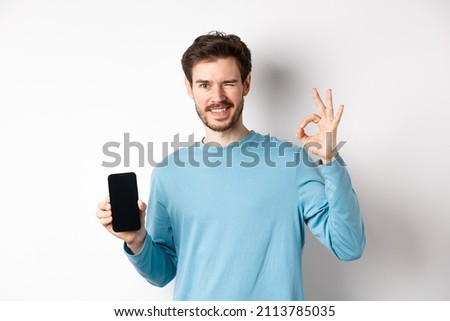 E-commerce and shopping concept. Smiling guy wink and show okay sign with empty smartphone screen, recommending online offer, standing on white background