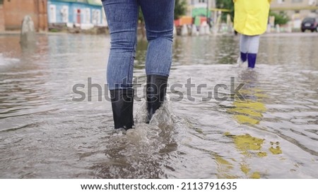 two people walk through puddles of flooded city rubber boots on their feet, teamwork, girls summer raincoats, lot of rainwater is poured on street, natural weather phenomena, walking boots on road Royalty-Free Stock Photo #2113791635