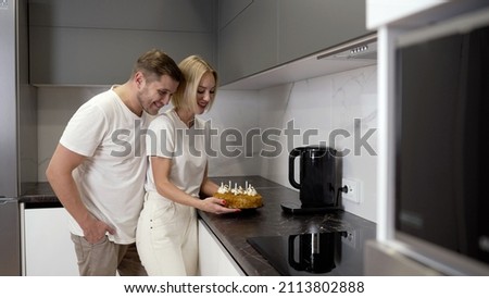 A guy with a girl and a cake. Young surprised girl holding a birthday cake and her boyfriend hugging her from behind. Young parents at home in the kitchen with cake.