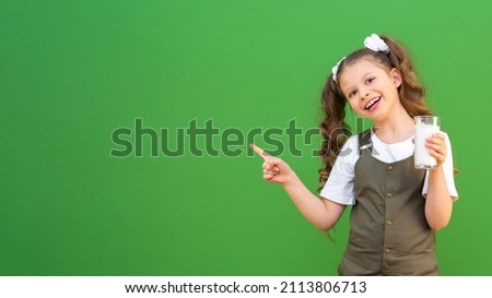 A little girl drinks cow's milk and points her finger at an advertisement on an isolated green background. Calcium for the child's body, copy space.