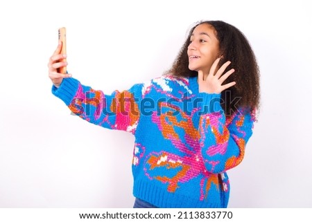 beautiful teenager girl wearing colorful sweater over white background holds modern mobile phone and makes video call waves palm in hello gesture. People modern technology concept