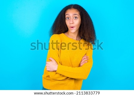 Shocked embarrassed beautiful teenager girl wearing yellow sweater over blue background keeps mouth widely opened. Hears unbelievable novelty stares in stupor