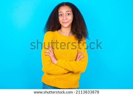 Self confident serious calm beautiful teenager girl wearing yellow sweater over blue background stands with arms folded. Shows professional vibe stands in assertive pose.
