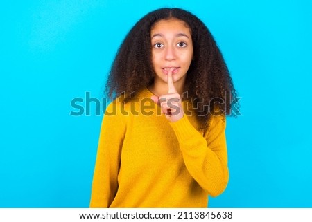Surprised beautiful teenager girl wearing yellow sweater over blue background makes silence gesture, keeps finger over lips and looks mysterious at camera