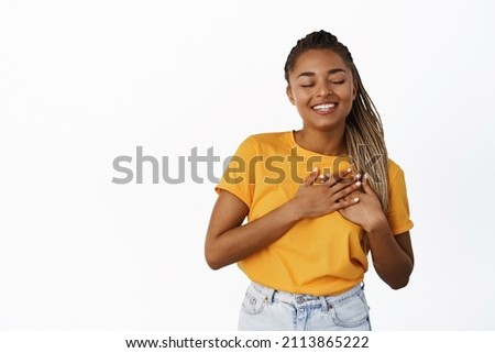 Image of beautiful smiling black woman close eyes and smile while holding hands on heart, thinking of something,having memory, standing over white background