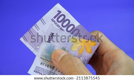 A man's hand is making a payment. Male hand showing Indonesian rupiah note. Indonesian Rupiah the official currency of Indonesia. Business concept, Blue Background. Uang 10000 Rupiah Bank Indonesia