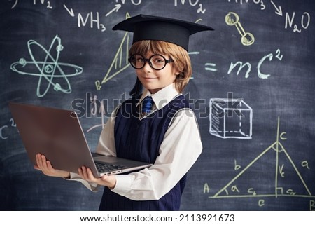 Portrait of a clever little boy in school uniform and glasses holding a laptop in the background of a blackboard with scientific formulas and diagrams. Smart children. Education.  Royalty-Free Stock Photo #2113921673