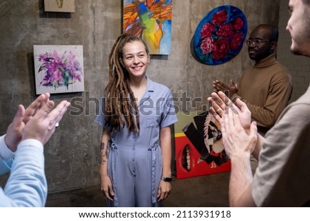 Successful young artist presenting her paintings in art gallery, she standing and smiling while people greeting her