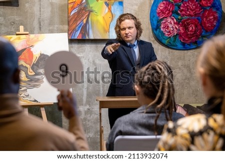 Mature man in suit pointing at man with tablet, people buying paintings of famous artists during auction in gallery